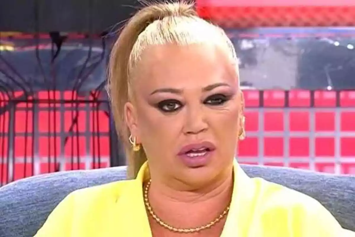 Una persona con expresión seria y maquillaje oscuro en un set de televisión con fondo rojo.