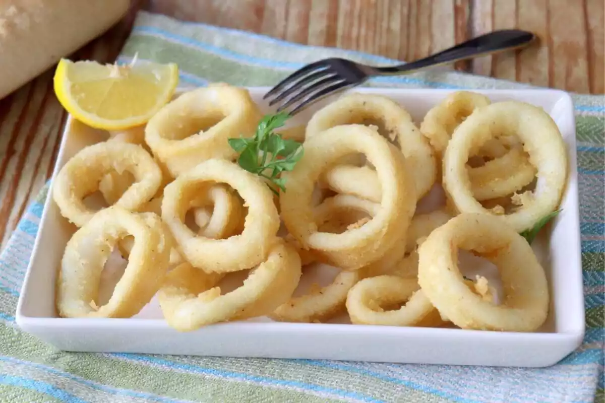 Un plato de calamares fritos con una rodaja de limón y un tenedor sobre un mantel a rayas.