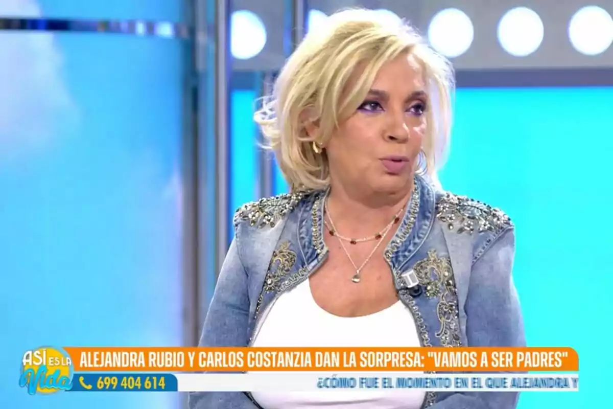 Una mujer con cabello rubio y chaqueta de mezclilla decorada aparece en un programa de televisión con un fondo azul y luces circulares.