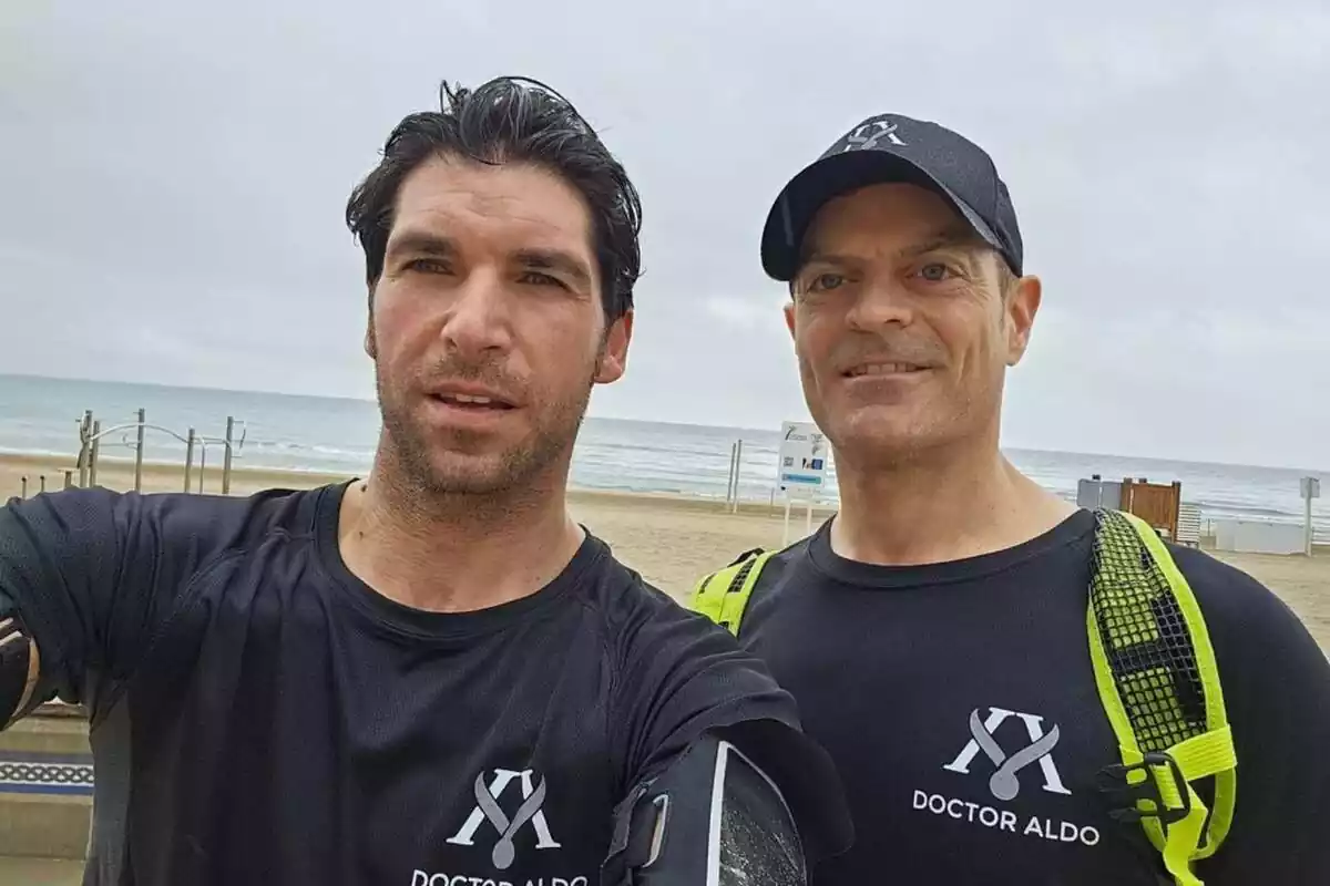 Dos hombres con camisetas negras que dicen "Doctor Aldo" posan para una foto en la playa con el mar de fondo.