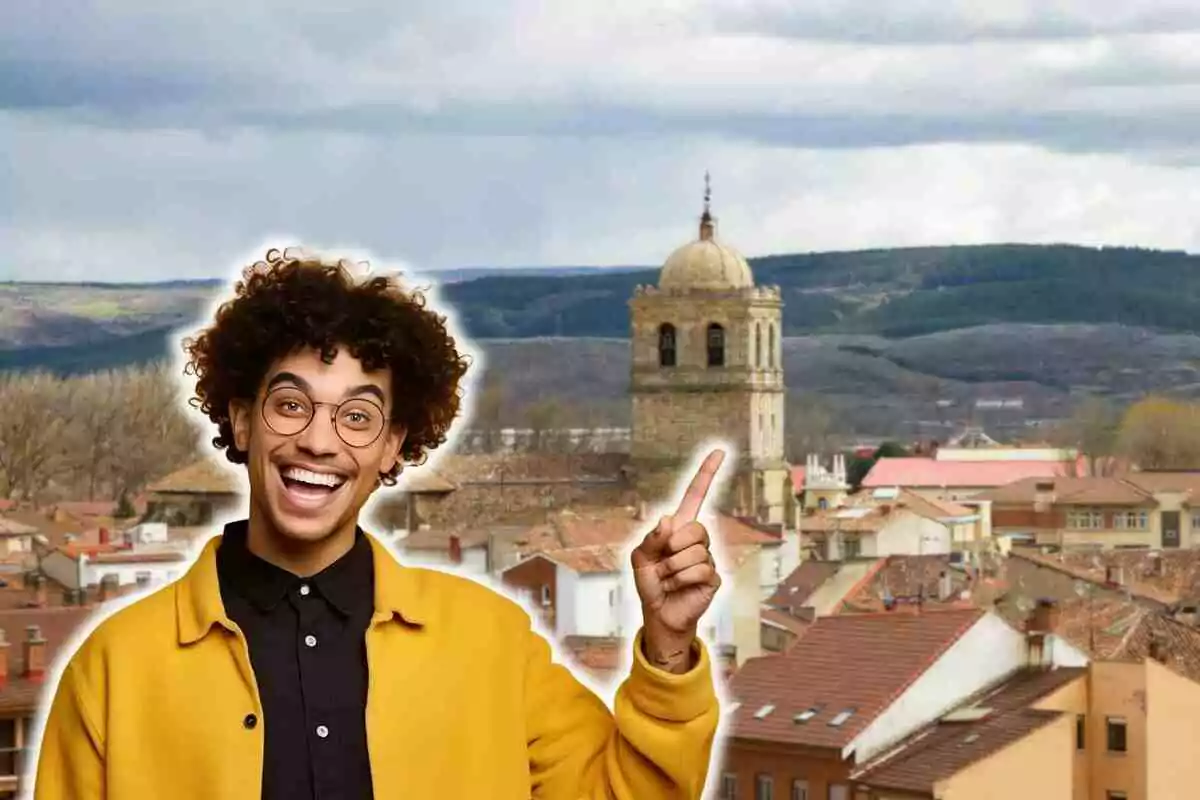 Un hombre sonriente con gafas y chaqueta amarilla señala hacia arriba con el dedo índice, con un paisaje urbano y una torre de iglesia al fondo.