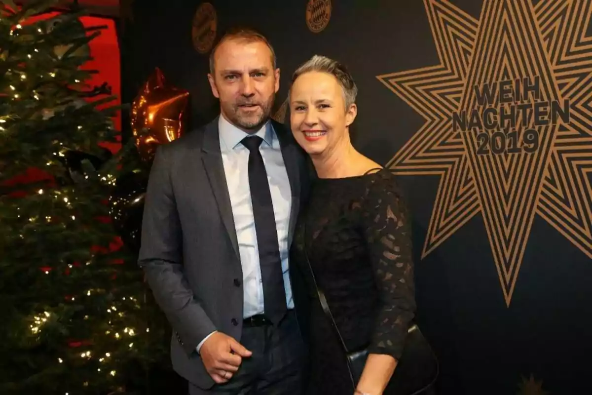 Una pareja sonriente posa frente a un árbol de Navidad decorado y un fondo con un diseño estrellado que dice "Weihnachten 2019".