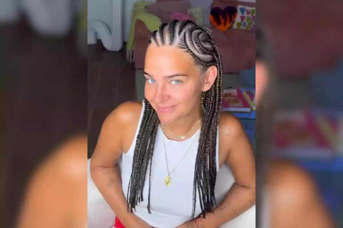 Una mujer con trenzas africanas y una camiseta blanca sonríe mientras está sentada en una habitación decorada con cojines coloridos y una manta verde.