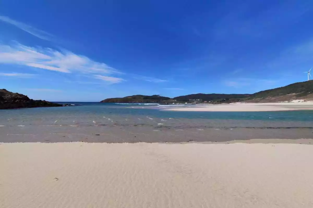 Playa de arena blanca con agua azul clara y colinas verdes al fondo bajo un cielo despejado.